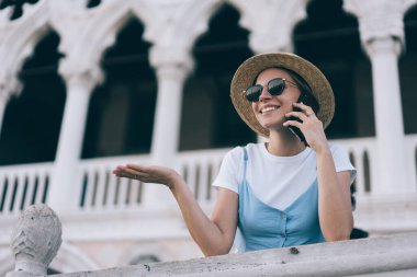 Moda kıyafetli mutlu beyaz kadın turist akıllı telefondan uluslararası arama yapıyor, güneş gözlüklü genç kadın yeni cep telefonu uygulaması aracılığıyla konuştuğu için en iyi arkadaşıyla iletişim kuruyor.