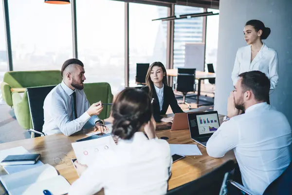 stock image Male employee sitting with colleagues at desk with gadgets and laptop with charts and talking to standing female manager during meeting in office with glass walls