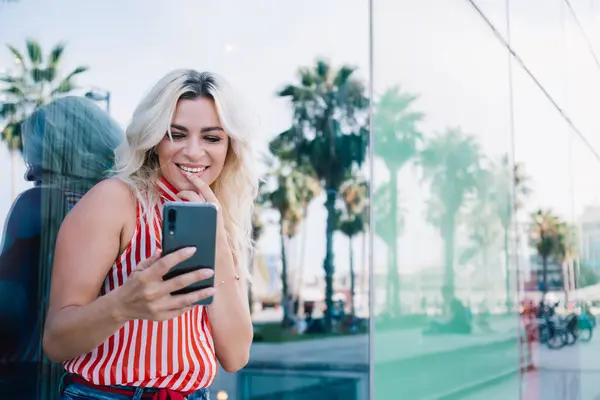 Young fair haired lady in striped tank top looking at phone screen and laughing happily with finger on chin while taking selfie on cellphone
