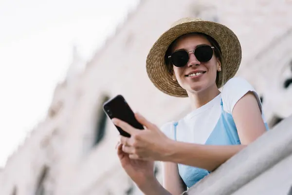 Portrait of female traveller in straw hat and trendy sunglasses holding mobile technology in hands and smiling during wireless phoning with roaming internet, happy tourist enjoying summer vacations