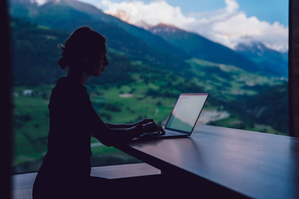 Вид сбоку на опытного кавказского цифрового кочевника, работающего дистанционно над фрилансным проектом, сидящего за столом возле большого окна с живописным видом на высокие горы, женщина тысячелетия печатает текст контента на ноутбуке