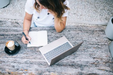 Kafede kahve ve dizüstü bilgisayarla otururken akıllı telefondan konuşan ve not defterine yazan bir kadından.