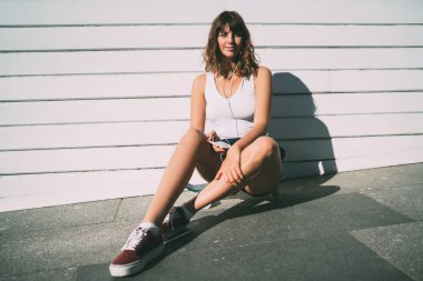 Genç bir bayan, günlük giysiler içinde kulaklıkla müzik dinliyor ve kaykay yaparken kameraya bakıyor ve beyaz duvarın yanındaki kaldırımda dinleniyor.