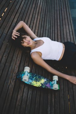 Beyaz tişörtlü ve siyah etekli muhteşem bir kadının yüksek açısı. Şehir parkındaki canlı kaykayın yanında kahverengi ahşap bankta yatarken kameraya gülümsüyor.