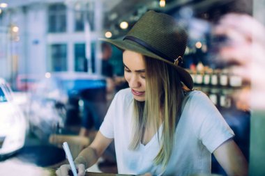 Camdan manzaralı, tropikal şapkalı ve beyaz gömlekli genç bir kız, arka planda bulanık bir kafede otururken yazı yazıyor.