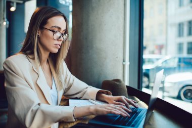 Kentteki kafede uzaktan çalışırken dizüstü bilgisayarda yazan bej takım elbiseli ve gözlüklü ciddi bir serbest çalışan genç bayan.