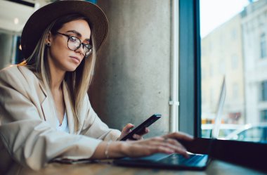 Gözlüklü, şapkalı, telefonu elinde tutan ve kafenin penceresine yakın bir yerde dizüstü bilgisayar kullanan düşünceli, zarif bir kadının yan görüntüsü.