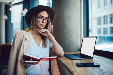 Zarif genç serbest çalışan elinde bir defter tutarak kafeterya masasının yanında boş ekranlı bir şekilde otururken düşünüyor.