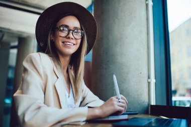 Zarif şapkalı ve gözlüklü güzel bir bayan elinde kalem ve kameraya gülümserken kafe masasında dizüstü bilgisayarının yanında oturuyor.