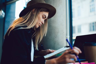 Restoranda sınava hazırlanırken elinde kalem tutan ve panodan veri okuyan şapkalı genç bir kadının yan görüntüsü