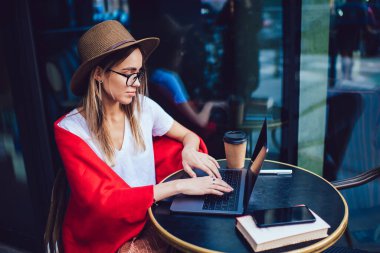 Kafe masasında otururken dizüstü bilgisayarında yazan, kendine güvenen, yaz şapkalı ve gözlüklü sarışın kadının yan görüntüsü.