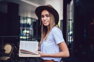 Neşeli, şapkalı, sıradan kıyafetler giyen, kitap okuyan, cam duvarın yanında durup kameraya gülümseyen güzel bir kadın.