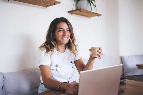 Joyful laughing young woman browsing internet on laptop and watching funny content while sitting at table with cup of coffee