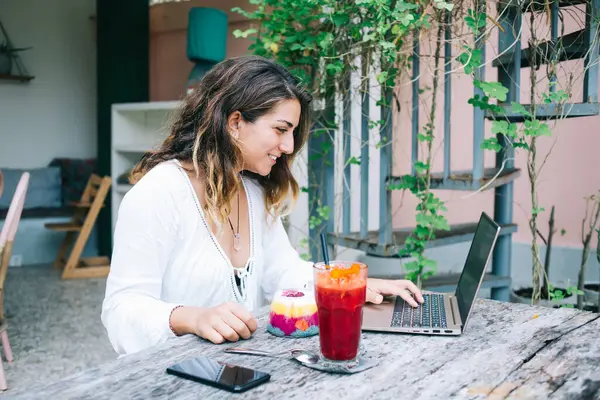 Side view of energetic and confident woman typing on keyword and eating berry bright juicy jelly with spoon sitting at table with red cocktail in outside cafe in yard