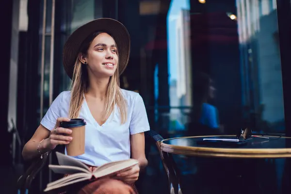 在城市咖啡店里 穿着时髦衣服 头戴帽子的无忧无虑的女人坐在桌子旁看书 喝咖啡 — 图库照片