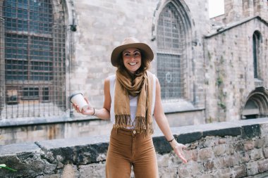 Tek kullanımlık bardakla gülümseyen genç bir kadın eski binanın yanında duruyor ve yürümek için yön seçerken ellerini açıyor.