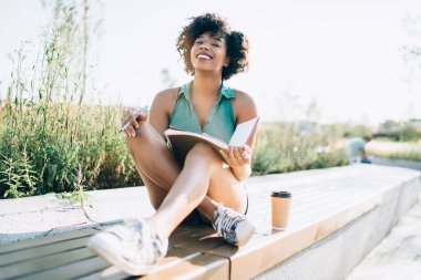 Güneşli bir günde şehir parkını öğrenmekten keyif alan zengin, esmer tenli bayan öğrenci, Afro-Amerikan kadın blogcu not defterinde yayınlanma fikirleri üzerine notlar alıyor.