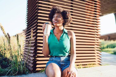 Neşeli, koyu tenli, kahve içmeyi seven, şehir parkında eğlenen, modaya uygun giyinmiş, Afro-Amerikan hippi kız güneşli bir günde dışarıda oturmuş içecek içiyor.