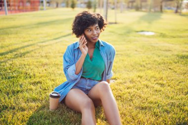 Güzel koyu tenli hippi kız cep telefonuyla konuşuyor yeşil çimlerde oturuyor açık havada eğleniyor, Afrika kökenli Amerikalı kadın milenyum yeşil çimlerde oturuyor ve iyi tarifeler ve cep telefonlarıyla konuşuyor.