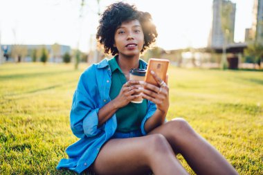 Genç Afro-Amerikalı kadın kot gömlek giyiyor ve parkta çimlerin üzerinde oturuyor. Yazın elinde kahve ve akıllı telefon varken.