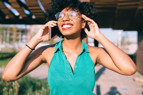 Cheerful dark skinned woman in trendy spectacles happy about free calls in roaming talking on mobile phone,happy emotional dark skinned female with curly hair laughing during smartphone talk