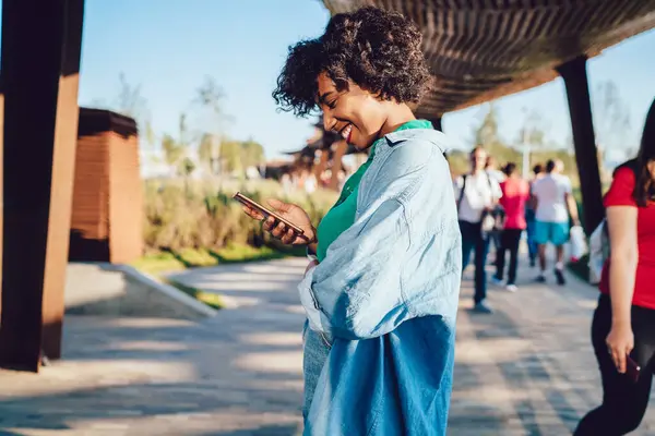 Side view of smiling black woman standing on covered walkway in park and reading pleasant text message on smartphone on blurred background with strolling people