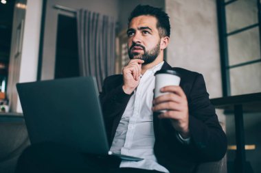 Resmi olarak ciddi düşünceli erkek girişimci internetteki iş bilgilerini analiz ederken ve elinde tek kullanımlık kahveyle dizüstü bilgisayarın önünde otururken karar verirken düşünüyor.