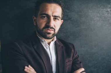 Karizmatik, zarif, resmi takım elbiseli, kollarını kavuşturmuş, iş yerinde kameraya bakarak vakit geçiren etnik bir adamın yakın çekimi.