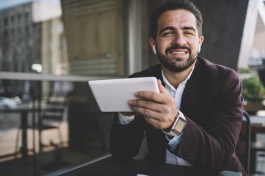 Modaya uygun giyinmiş, gülümseyen ve kameraya bakan iyimser etnik adam balkonda güzel müziğin ve modern tabletin tadını çıkarıyor.