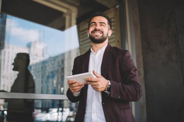 Modern binanın penceresinin yanında müzik dinlerken ve tablet kullanırken gülümseyen, bakışlarını çeviren modaya uygun giyinmiş mutlu etnik yöneticinin düşük açısı