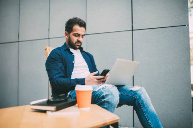 Düşük açılı, ciddi düşünceli serbest çalışan, günlük kıyafetlerle ekranlara odaklanan ve akıllı telefonla iletişim kuran modern kafedeki bulanık gri duvara karşı koymak için ahşap masadaki netbook ve sıcak içecekle birlikte tahta masada oturan biri.