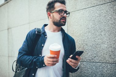 Alttaki mahsulden, sakin, düşünceli, gözlüklü ve sıcak içecekle sıradan giyimli serbest çalışan. Şehirdeki bulanık gri duvara karşı akıllı telefonla iletişim kurarken başka bir yere gitmek ve düşünmek için.