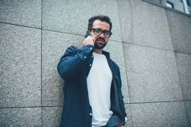 Gözlüklü, beyaz ve mavi tişörtlü, şık sakallı bir adam şehrin gri kareli duvarına karşı sokakta dikilirken gözlerini kaçırıp telefonla konuşuyor.