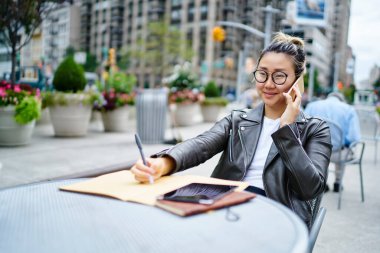 Deri ceketli Japon milenyumu ve vizyon koruması için klasik gözlükler. Arkadaşını buluşmayı planlamak için sokak kafeteryasında dolaşma bağlantısını kullanıyor. Uygulamayla arayan Asyalı kadın.