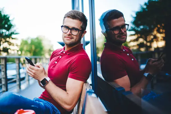 Portrait of handsome caucasian hipster guy in stylish eyewear phoning while having break outdoors, prosperous blonde young man chatting via smartphone application use gadgets sitting on street