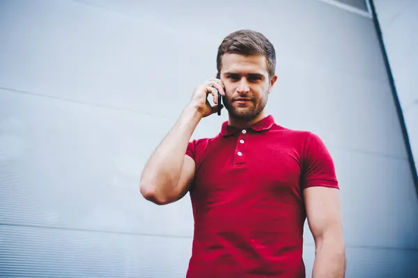Handsome caucasian male in pink t-shirt talking on mobile phone in roaming looking at camera, portrait of young 20s hipster guy satisfied with connection making smartphone call posing on mock up wall