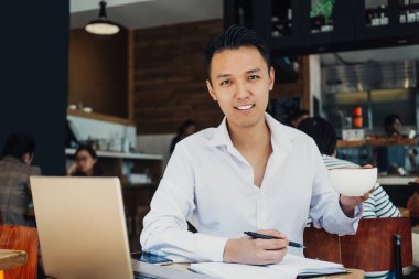 Zarif beyaz gömlekli, elinde kahve fincanıyla kafede oturan ve kameraya gülümseyen genç Asyalı adam. 