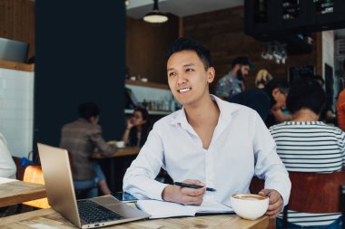 Beyaz tişörtlü, laptoplu ve not defterli modern gülümseyen Asyalı adam kahve içerken yüzünü çeviriyor. 