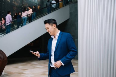 İş adamı kılığındaki Asyalı genç adam gülümsüyor ve iş merkezinde yürüyen merdivene karşı dururken akıllı telefondan mesajlaşıyor.