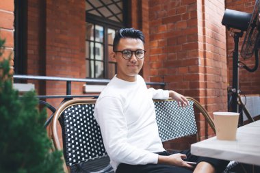 Tatmin olmuş gözlüklü genç erkek öğrenci kameraya bakıyor ve terasta oturup dinlenirken telefonu tutuyor.