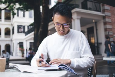 Kafe dışındaki masada ders çalışırken ve marj notları alırken odaklanmış, Asyalı stil sahibi bir öğrenci kitabı okuyor. 