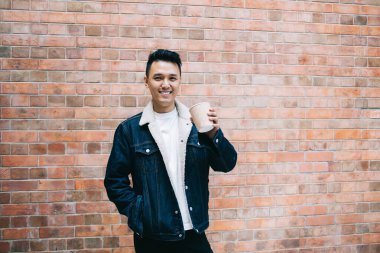 Koyu saçlı Asyalı, kot ceketli, beyaz tişörtlü, dişlek, plastik bardak tutan ve kameraya bakarken kahve içen.