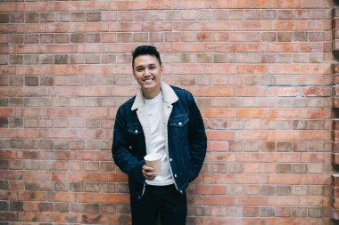 Kahveli Asyalı güler yüzlü genç adam. Cebinde tuğla duvarın yanında dikilip kameraya bakıyor.