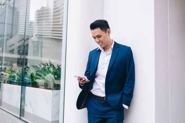 Asian young man in business suit leaning on white wall near glass fence and smiling while texting on smartphone on city street