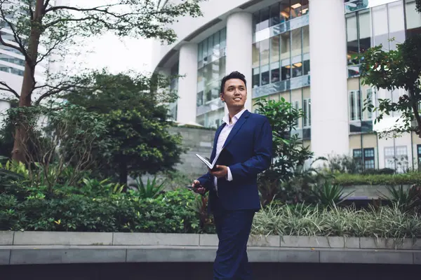 Handsome adult worker in formal clothes standing outdoors against green fence and contemporary building holding phone and planner smiling looking away