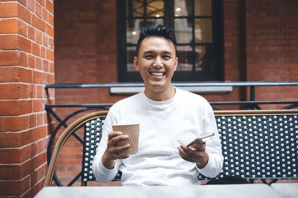 Cheerful youthful dark haired Asian man looking at camera and using smartphone while holding takeaway coffee and sitting at table outdoors