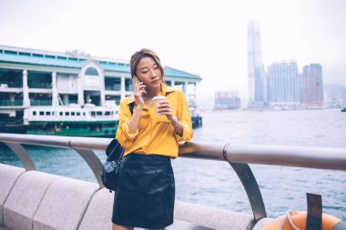 Sarı tişörtlü, siyah deri etekli çekici bir Asyalı kadın kahve alıp Hong Kong 'da resim gibi bir feribot iskelesinde telefonla konuşuyor.