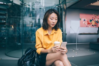 Şık etekli, sarı tişörtlü düşünceli Asyalı kadın modern iş merkezinde sırt çantasıyla oturmuş kahve tutuyor ve cep telefonlarını karıştırıyor.