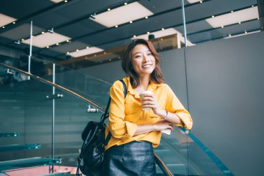 Çekici, neşeli, sarı gömlekli, siyah deri etekli, sırt çantalı, sıcak içecek ve cep telefonlu güzel bir Asyalı kadın Hong Kong 'da kapalı kapılar ardında duruyor.