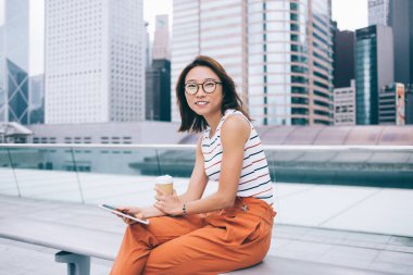 Çekici, modaya uygun kıyafetleri ve bardakları olan, elinde tek kullanımlık kahve ve tabletle Hong Kong 'da bir bankta oturmuş kameraya bakan Asyalı bir kadın. 